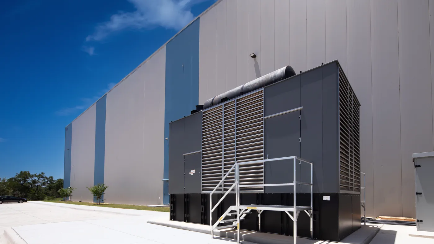 A large commercial standby generator with an access platform, standing against the wall of a metal-clad warehouse